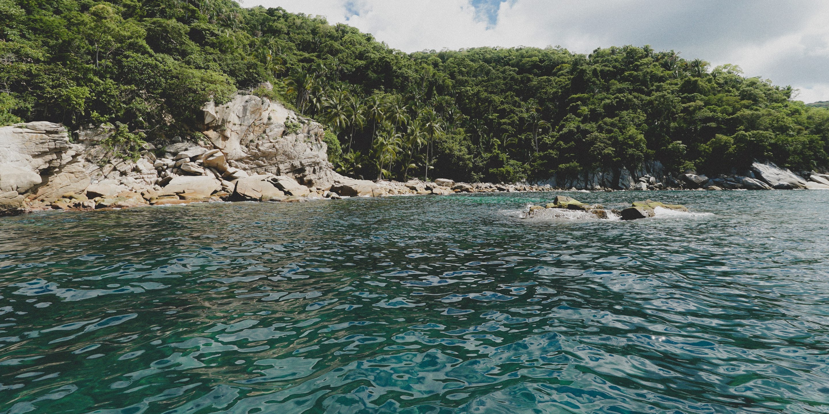 Costa de roca y selva vista desde el agua, con el mar turquesa al frente.